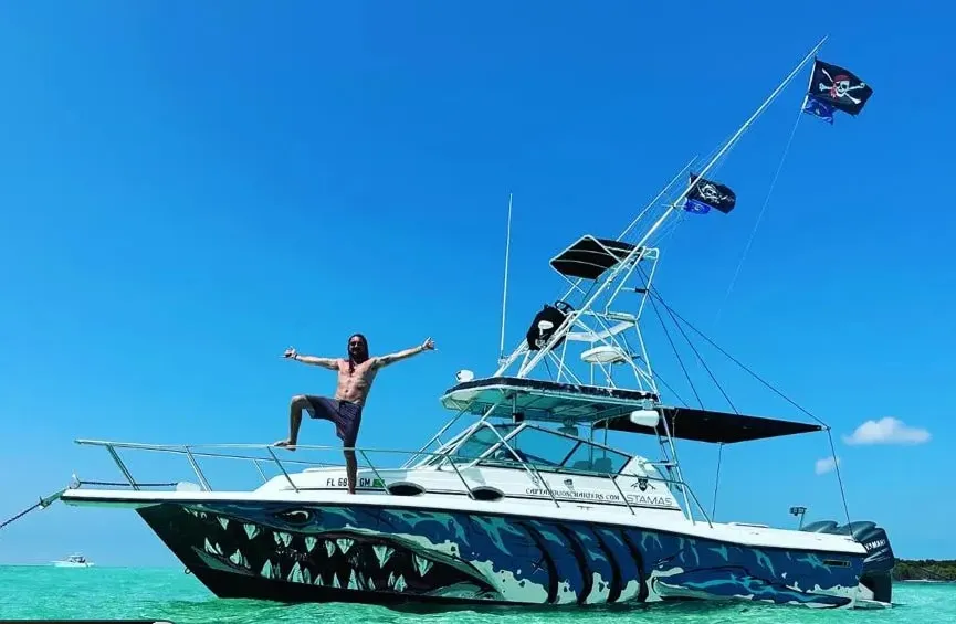 Captain Rio standing on the bow of his shark-mouth painted 32 ft Stamas, anchored over a turquoise Key West sandbar with pirate flags flying